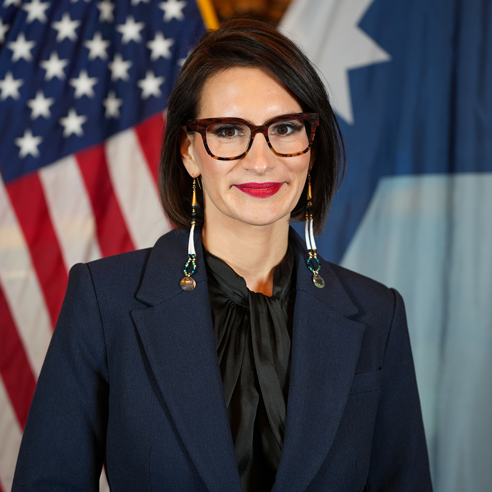 Headshot of Lt. Governor Flanagan wearing a dark blue blazer, black silk shirt, long beaded earrings, and dark-rimmed glasses